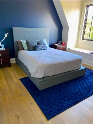 A clean and simple solid wood platform bed with a grey upholstered headboard in a bedroom.